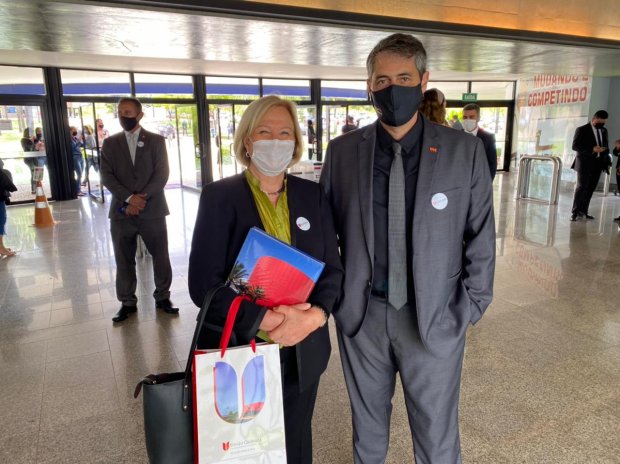 Duas pessoas posam para foto em ambiente interno, ambas usando máscara. À esquerda, uma mulher segura sacolas e uma pasta azul com estampa vermelha. À direita, um homem de terno cinza escuro está com as mãos nos bolsos. Ao fundo, outras pessoas de máscara também circulam pelo local, que tem portas de vidro e piso claro. No canto direito da imagem, vê-se parcialmente a frase 'Mundos Competindo' na parede.
