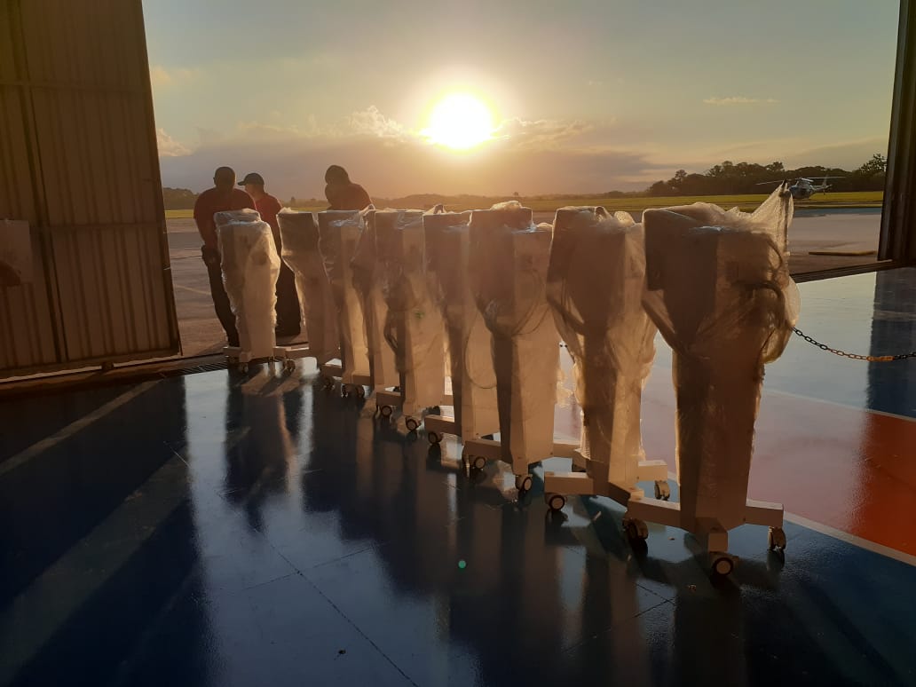 Fileira de ventiladores pulmonares embalados em plástico, posicionados na entrada de um hangar durante o pôr do sol. Ao fundo, três pessoas observam o cenário na pista do aeroporto.