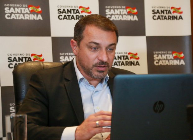 Homem usando terno escuro e camisa clara participa de videoconferência em frente a um notebook HP. Ao fundo, painel com o logotipo do Governo de Santa Catarina, exibido em padrão quadriculado preto e branco, com a bandeira do estado.