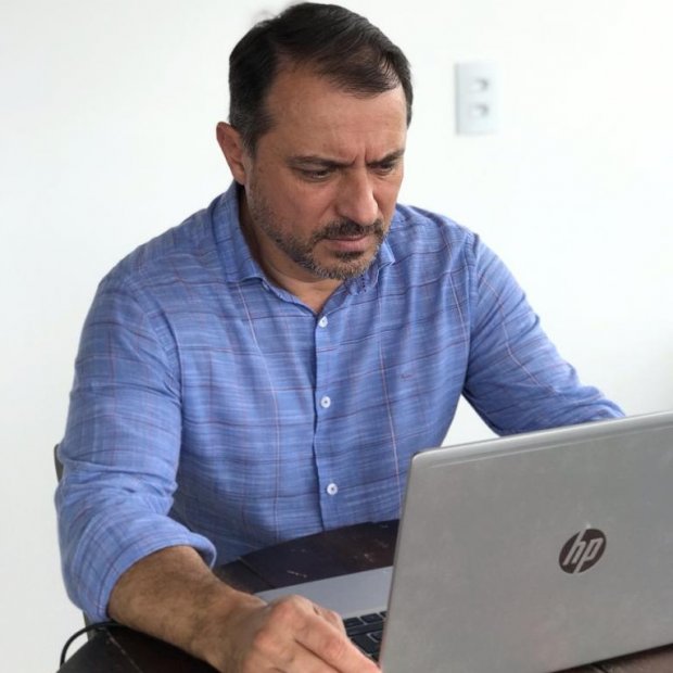 Homem de meia-idade, com barba por fazer e camisa azul, está sentado em frente a um notebook da marca HP. Ele aparenta estar concentrado enquanto trabalha. Ao fundo, há uma parede branca com uma tomada visível.