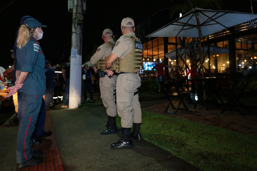 Agentes da Polícia Militar e da Vigilância Sanitária realizam fiscalização noturna em frente a um restaurante ou bar iluminado. Os policiais estão de costas, observando o movimento no local, que tem mesas externas, guarda-sóis e clientes sentados. Cena mostra atuação das autoridades no controle de aglomerações durante a pandemia.