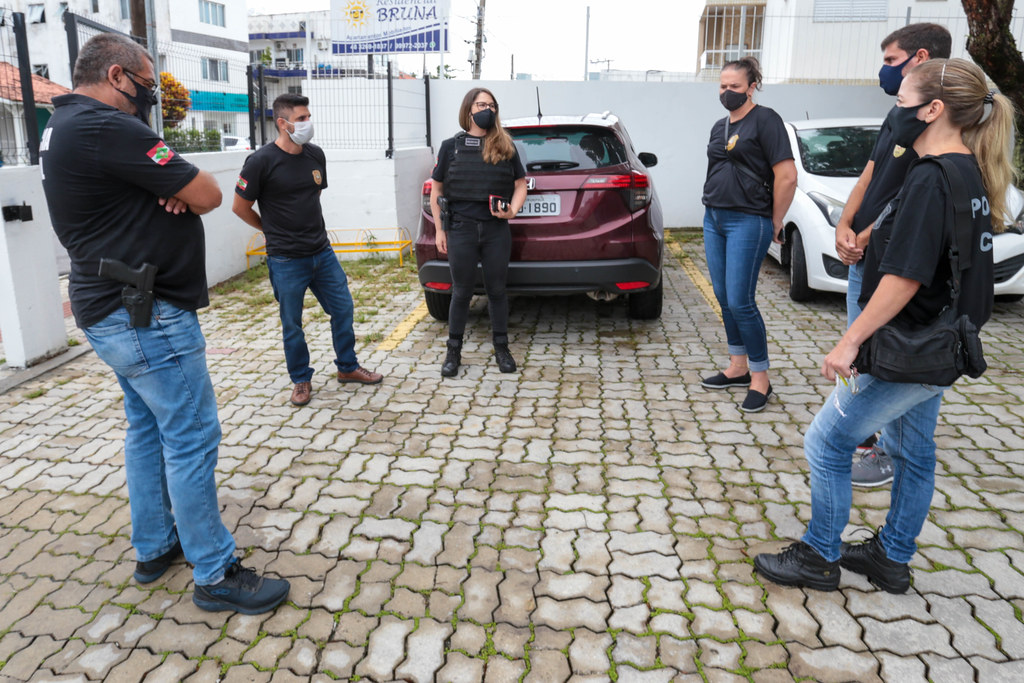 Grupo de agentes da Polícia Civil de Santa Catarina, com coletes e camisetas identificadas, realiza reunião ao ar livre em um estacionamento com piso de blocos intertravados. Eles estão organizados em semicírculo, próximos a veículos estacionados, incluindo um carro vermelho ao centro. Todos utilizam máscaras de proteção, indicando contexto de pandemia.