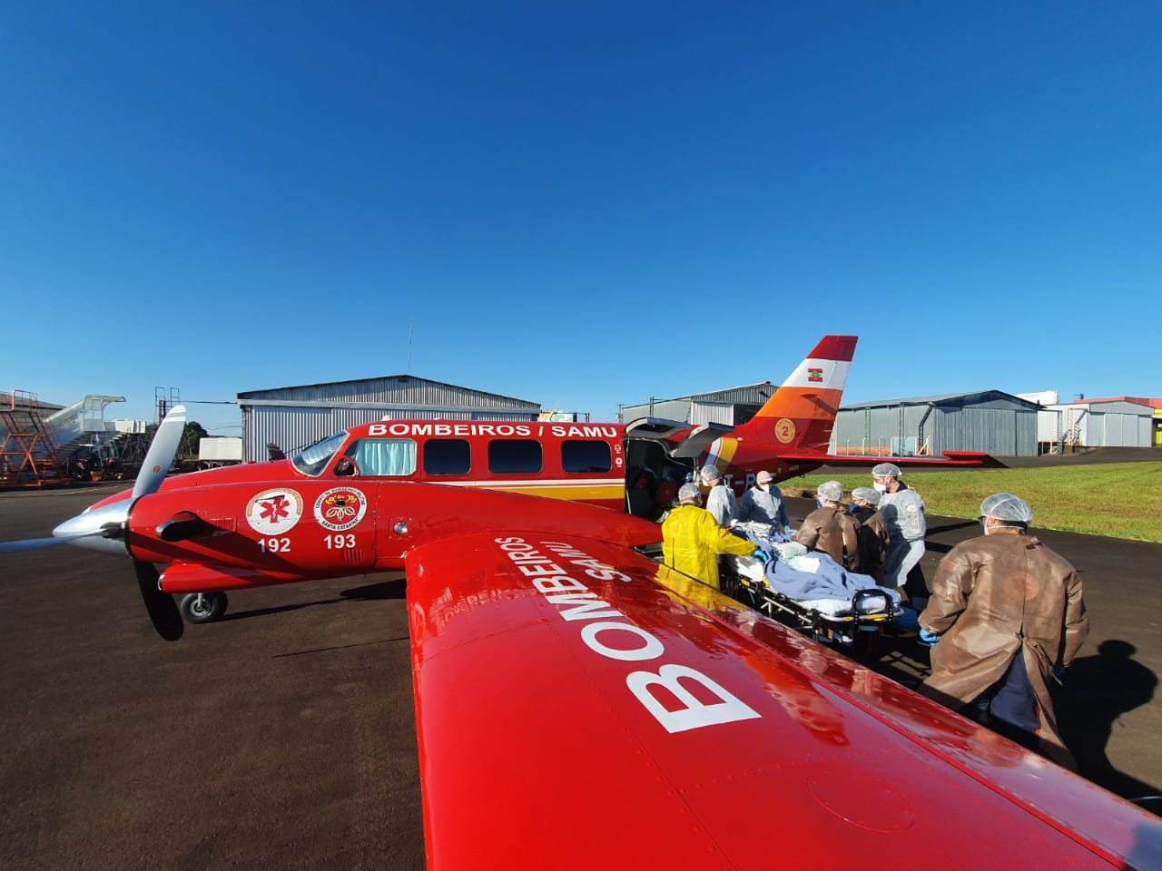 Equipe do SAMU e Bombeiros realiza transporte aeromédico de paciente em maca até aeronave vermelha com inscrições BOMBEIROS / SAMU. A ação ocorre em aeroporto sob céu azul e tempo aberto.