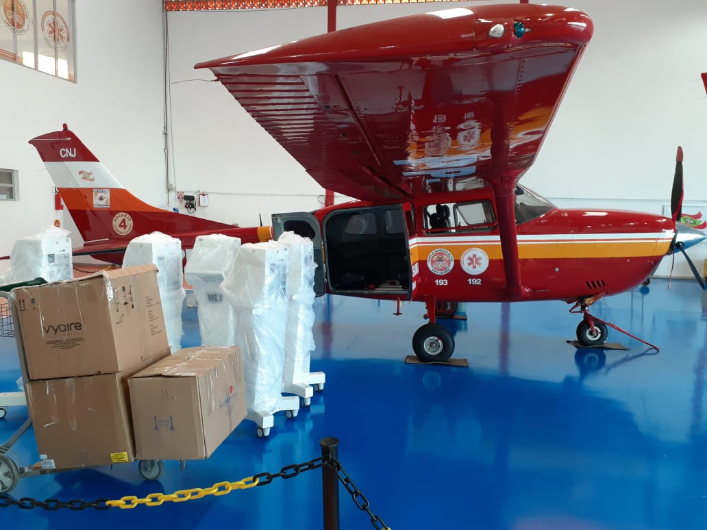 Avião vermelho do Corpo de Bombeiros de Santa Catarina estacionado em um hangar, com a porta lateral aberta. Ao lado da aeronave, há caixas de papelão e equipamentos médicos embalados em plástico, prontos para transporte. No chão azul brilhante do hangar, a cena indica uma operação logística de envio de suprimentos, possivelmente relacionados à saúde.