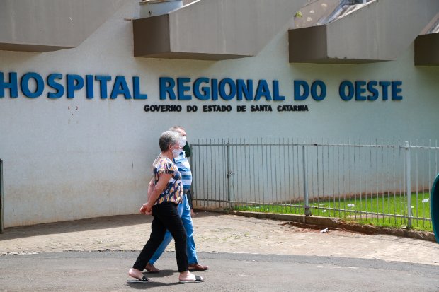 Duas pessoas com máscaras caminham em frente ao Hospital Regional do Oeste, em Santa Catarina, com o nome do hospital visível na fachada.