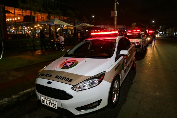 Viatura da Polícia Militar de Santa Catarina com giroflex ligado estacionada em frente a um restaurante iluminado à noite. Outras viaturas estão enfileiradas atrás. Operação policial em área urbana.