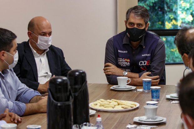 Reunião com seis homens sentados ao redor de uma mesa de madeira, todos usando máscaras. No centro da mesa, há garrafas térmicas, copos, xícaras e dois pratos com biscoitos. Um dos participantes usa colete com o logo do Governo de Santa Catarina e COES (Centro de Operações de Emergência em Saúde). Ao fundo, há uma janela com vista para árvores.