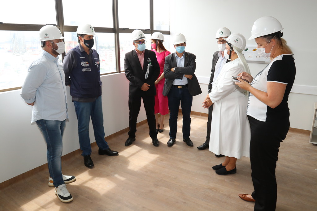 Grupo de pessoas usando capacetes de proteção e máscaras realiza visita técnica em ambiente interno iluminado, com janelas amplas. Entre os visitantes, há profissionais da saúde e representantes do governo.
