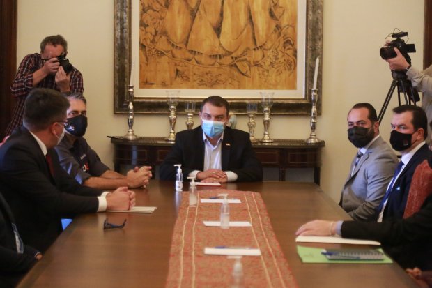 Reunião formal com oito homens sentados ao redor de uma mesa de madeira longa, todos usando máscara facial. Ao centro da mesa há frascos de álcool em gel e papéis. Ao fundo, uma parede com uma pintura grande e candelabros. Dois fotógrafos registram o momento de pé nos cantos da sala. A cena sugere um encontro governamental ou institucional.
