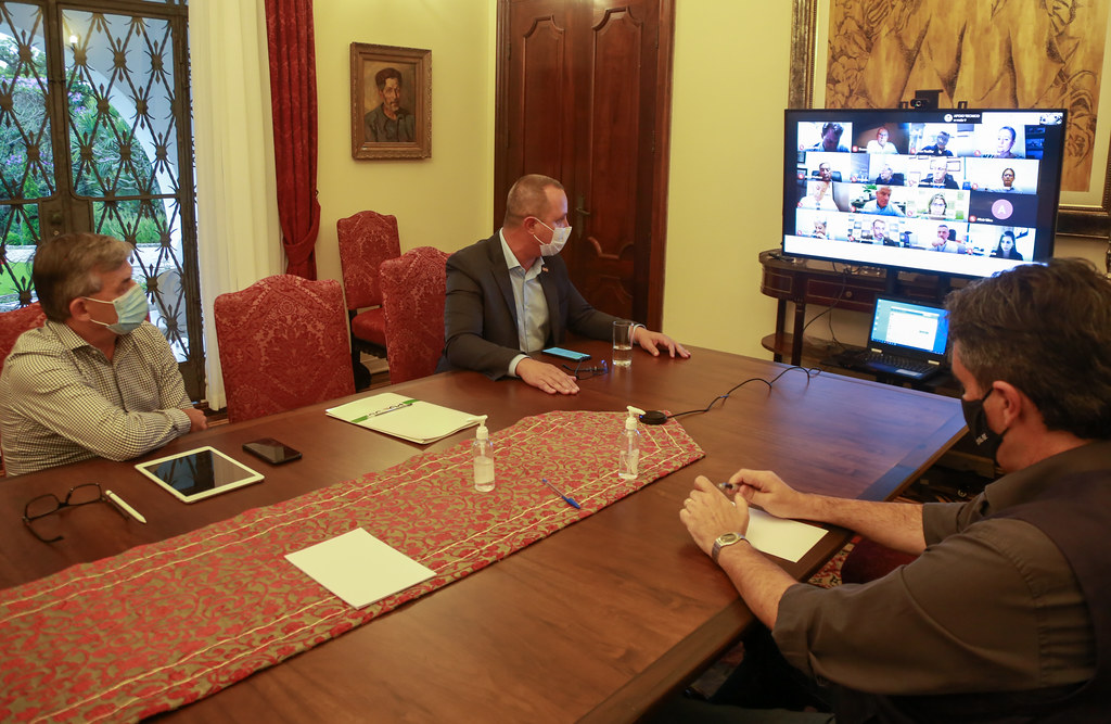 Três homens usando máscaras participam de uma reunião presencial em torno de uma mesa de madeira, enquanto interagem com outras pessoas por videoconferência, exibida em uma grande tela à frente. Sobre a mesa, há álcool em gel, papéis, eletrônicos e cadernos. O ambiente tem decoração clássica, com móveis antigos, cortinas vermelhas e uma pintura na parede.