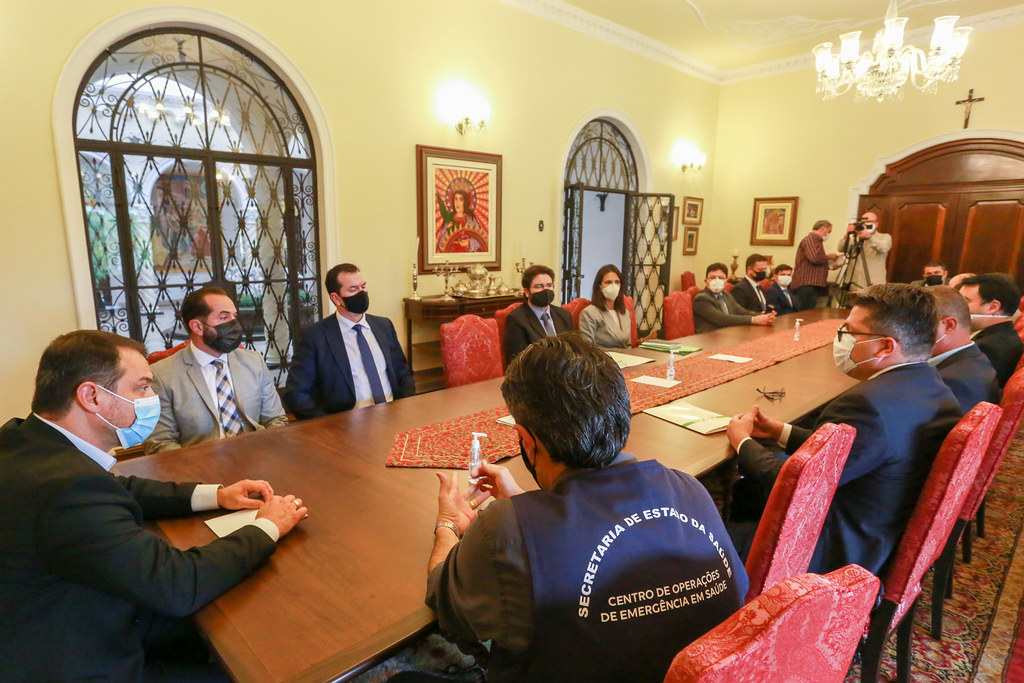 Reunião formal em uma sala elegante com paredes amarelas, lustre e móveis antigos. Cerca de 16 pessoas, todas usando máscaras, estão sentadas ao redor de uma mesa longa de madeira com cadeiras vermelhas. Um dos participantes usa colete escrito 'Secretaria de Estado da Saúde – Centro de Operações de Emergência em Saúde'. Ao fundo, há dois fotógrafos registrando o encontro. A atmosfera é solene e institucional.
