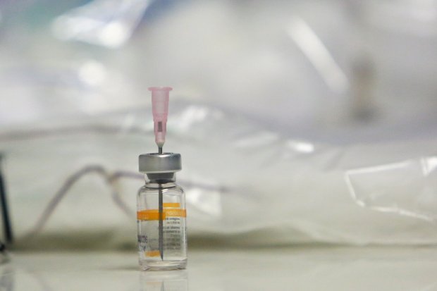Frasco de vacina sobre uma superfície branca, com uma seringa acoplada na parte superior. Ao fundo, há um plástico translúcido desfocado, sugerindo ambiente hospitalar ou laboratório. A imagem enfatiza o frasco em primeiro plano, remetendo à vacinação.