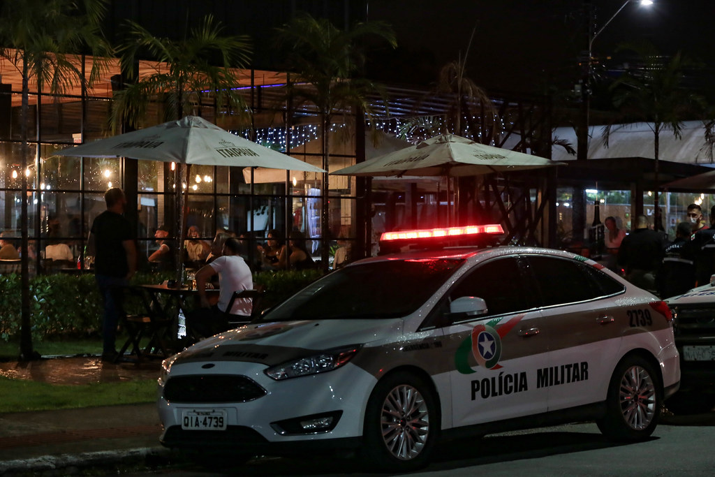 Viatura da Polícia Militar com giroflex ligado estacionada em frente a um bar ou restaurante movimentado durante a noite. O local tem grandes janelas de vidro, guarda-sóis com a marca Itaipava e mesas ocupadas por clientes. A cena é iluminada por luzes decorativas e postes de rua, com presença de policiais e frequentadores no ambiente externo.