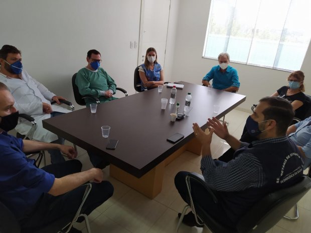 Reunião em uma sala com nove pessoas sentadas ao redor de uma mesa retangular, todas usando máscaras. Uma das pessoas, vestindo colete da Secretaria de Estado da Saúde, fala enquanto os demais escutam. A sala é clara, com parede branca e janelas ao fundo.
