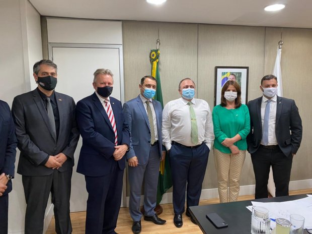 Grupo de sete pessoas usando máscaras posando para foto em sala oficial. Ao fundo, há as bandeiras do Brasil e de uma entidade não identificada, além de um retrato institucional. Todos estão vestidos com roupas formais. A cena se passa em um ambiente de reuniões, com mesa e copos de vidro em primeiro plano.