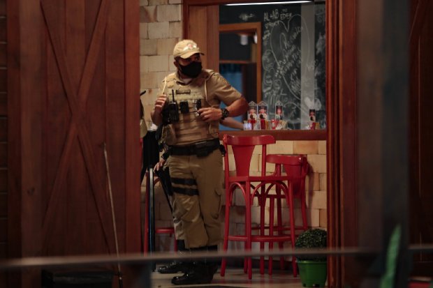 Policial militar com máscara e colete tático realiza fiscalização em estabelecimento comercial com cadeiras vermelhas e decoração de Natal.