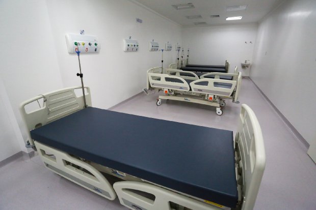 Sala hospitalar vazia com seis leitos equipados, posicionados em fileiras paralelas. As camas têm estrutura ajustável, colchões azuis escuros e estão conectadas a painéis com saídas de oxigênio e outros dispositivos médicos fixados na parede. O ambiente é limpo, bem iluminado e possui piso claro, refletindo um espaço preparado para receber pacientes.