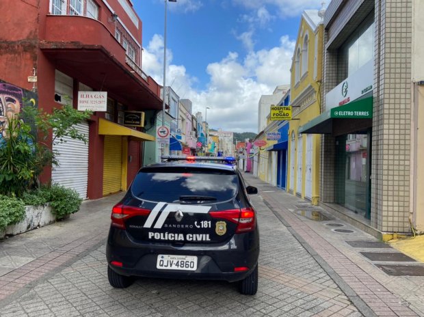 Viatura da Polícia Civil com giroflex ligado estacionada em uma rua de calçadão comercial, onde as lojas estão com as portas fechadas. A rua é ladeada por prédios coloridos e comerciais. O céu está azul com algumas nuvens, indicando dia claro e ensolarado.