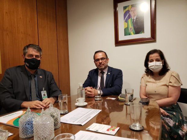 Três pessoas sentadas à mesa durante reunião, duas delas usando máscara; ao fundo, retrato oficial de autoridade brasileira na parede.