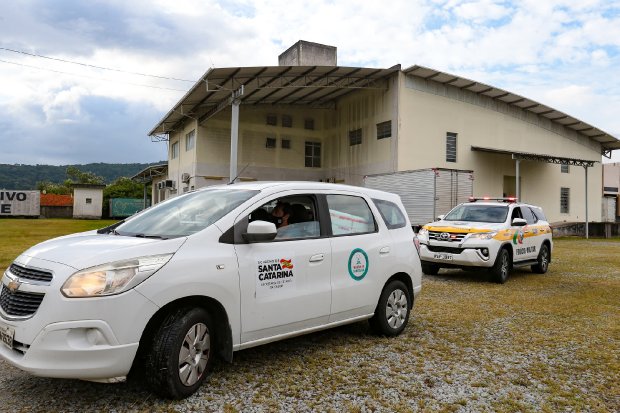 Carro branco oficial com o logotipo do Governo de Santa Catarina e da Secretaria de Saúde está estacionado em frente a um prédio de estrutura simples com telhado metálico. Atrás do veículo, uma caminhonete da Polícia Militar Rodoviária com luzes de emergência está parada. O local parece ser uma área de apoio ou centro logístico, com chão de brita e gramado. Ao fundo, há vegetação e céu parcialmente nublado. Cena relacionada à operação de transporte de vacinas ou insumos de saúde pública.