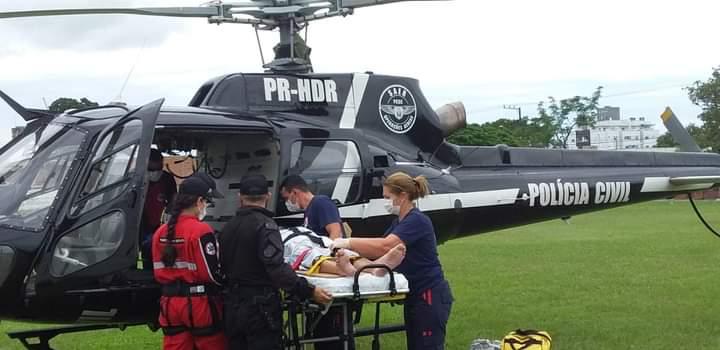 Equipe médica e policial realiza o transporte de um paciente em maca para um helicóptero da Polícia Civil com identificação PR-HDR, estacionado em um campo gramado. A cena indica uma operação de resgate ou transporte aeromédico de emergência.