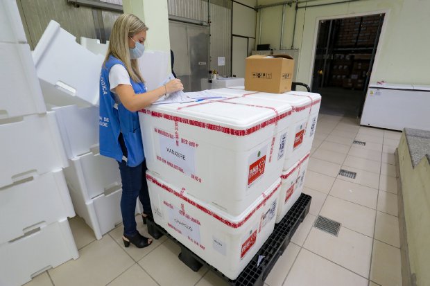 Profissional da saúde com colete azul da Secretaria da Saúde de Santa Catarina confere documentos ao lado de caixas térmicas rotuladas com o nome 'Xanxerê', indicando o destino das vacinas. As caixas estão sobre um pallet de plástico preto dentro de uma sala de armazenamento. A profissional usa máscara e está cercada por outras caixas térmicas e equipamentos de refrigeração. Cena relacionada à logística de distribuição de vacinas contra a COVID-19.