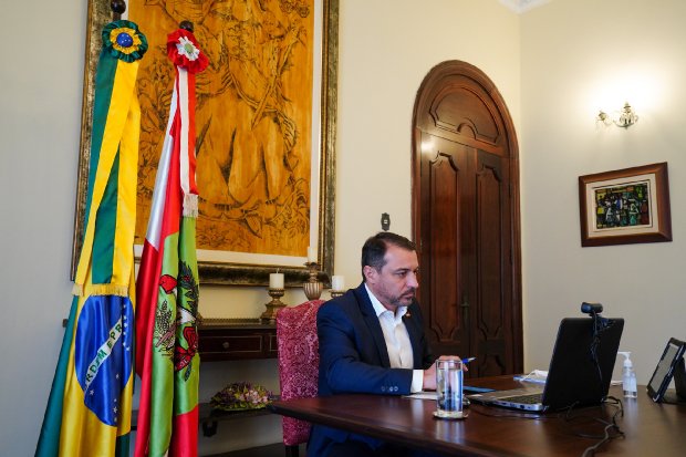 Homem de terno participa de reunião virtual em gabinete com bandeiras do Brasil e de Santa Catarina ao fundo.
