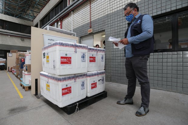 Homem com máscara e colete do Ministério da Saúde confere documentos ao lado de caixas térmicas brancas identificadas como frágeis, empilhadas sobre um pallet preto. A cena ocorre em área externa de um centro logístico com paredes de azulejo cinza, onde estão sendo recebidas vacinas ou insumos de saúde.