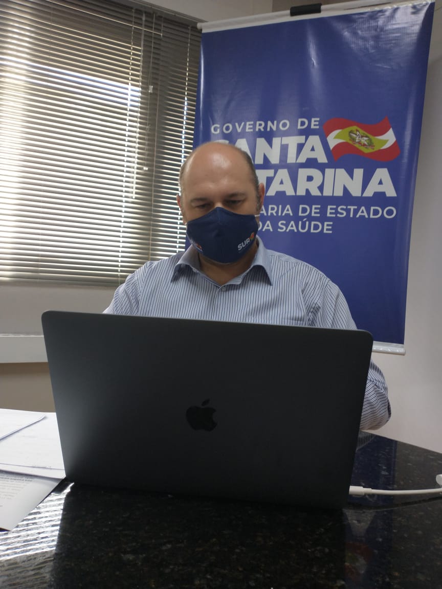 Homem calvo usando máscara facial azul com logotipo do SUS, sentado à mesa e trabalhando em um notebook da Apple. Ao fundo, há uma persiana e um banner do Governo de Santa Catarina com o texto Secretaria de Estado da Saúde.