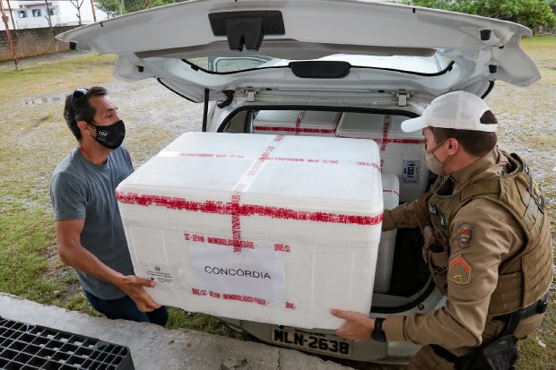Dois homens, um deles usando uniforme da Polícia Militar, retiram uma grande caixa térmica branca de um carro branco com a tampa do porta-malas aberta. A caixa está identificada com a palavra 'Concórdia' e lacrada com fitas vermelhas. Ambos utilizam máscaras faciais. A cena ocorre em área externa com grama e algumas árvores ao fundo. A placa do carro é 'MLN-2638'. A imagem retrata a logística de distribuição de vacinas ou insumos médicos durante a pandemia de COVID-19.