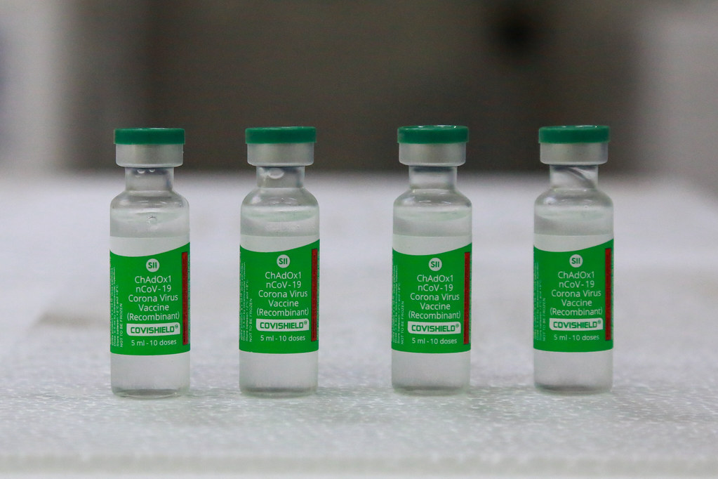 Quatro frascos da vacina Covishield (ChAdOx1 nCoV-19) estão alinhados sobre uma superfície branca. Os frascos possuem rótulo verde com detalhes em branco e vermelho, e tampas verdes. Cada frasco contém 5 ml, equivalentes a 10 doses da vacina contra a Covid-19, produzida pelo Serum Institute of India.