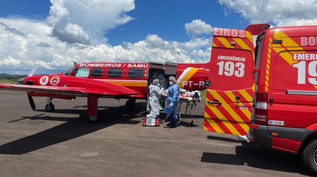 Equipe médica realiza transferência de paciente para avião vermelho do Corpo de Bombeiros e SAMU em uma pista de pouso, com ambulância de emergência 193 estacionada ao lado. Céu azul com nuvens ao fundo. Cena representa atendimento de urgência aeromédico em Santa Catarina.