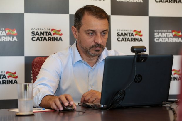 Homem de camisa azul clara participa de videoconferência usando um notebook HP com webcam acoplada. Ao fundo, painel com logomarcas do Governo de Santa Catarina. Sobre a mesa, copo com água e caneta.