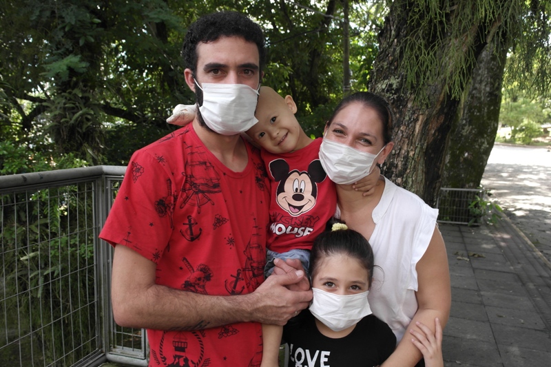 Família com quatro pessoas, incluindo duas crianças, posa ao ar livre usando máscaras de proteção. Um dos adultos segura no colo uma das crianças, que está vestida com camiseta do Mickey. Todos estão sorrindo e abraçados, em um ambiente com árvores e cercado por gradil.