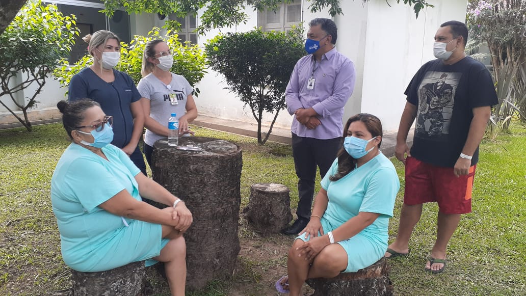 Seis pessoas com máscaras conversam ao ar livre em um jardim com árvores. Duas mulheres vestindo roupas hospitalares azul-claro estão sentadas em troncos de madeira. Outras quatro pessoas, incluindo profissionais de saúde com crachás, estão em pé ao redor. Ao centro, um tronco maior serve como mesa com uma garrafa de água sobre ele. Ao fundo, há um prédio branco.