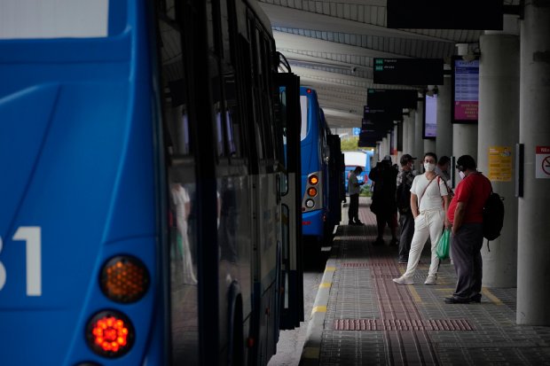 Pessoas usam máscaras enquanto aguardam na plataforma de um terminal de ônibus. Vários ônibus azuis estão estacionados lado a lado, e passageiros se preparam para embarcar. O local possui cobertura e sinalização suspensa.
