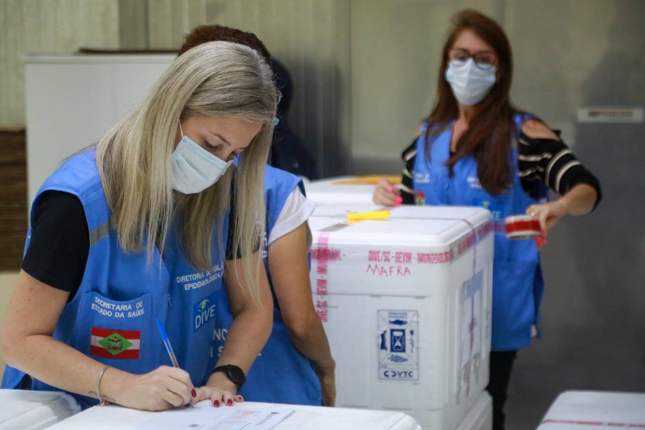 Profissionais da Diretoria de Vigilância Epidemiológica de Santa Catarina, usando coletes azuis e máscaras, conferem documentos e identificações em caixas térmicas rotuladas com nomes de municípios, como Mafra. A cena mostra o processo de controle e distribuição de vacinas contra a COVID-19 no estado.