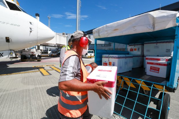 Trabalhador com colete laranja e protetor auricular descarrega caixa térmica com vacinas de uma carreta no pátio de aeroporto, próximo a um avião de carga. Ao fundo, mais caixas identificadas como contendo imunizantes. Cena sob céu azul e ensolarado.