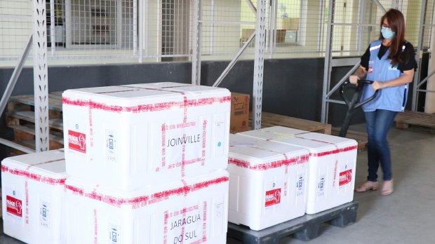 Servidora com máscara e colete azul manuseia paleteira com caixas térmicas identificadas para Joinville e Jaraguá do Sul, dentro de galpão logístico.