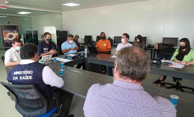 Reunião com várias pessoas sentadas ao redor de uma mesa em uma sala com computadores; todos usam máscaras. Um dos participantes veste colete com a inscrição (Ministério da Saúde). Há copos de água sobre a mesa e banner da Defesa Civil ao fundo.