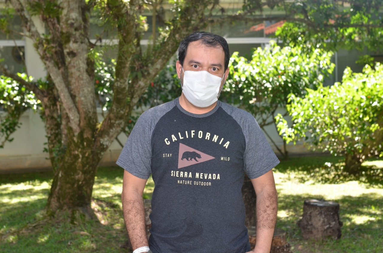 Homem em pé ao ar livre, usando máscara branca e camiseta cinza escura com a estampa (California Stay Wild Sierra Nevada Nature Outdoor), com árvores e vegetação ao fundo.