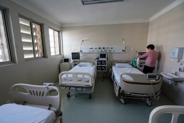 Quarto hospitalar amplo com quatro camas vazias, cada uma posicionada em um canto da sala. Uma profissional de saúde, vestindo uniforme rosa, está ao lado de uma das camas, avaliando o espaço.