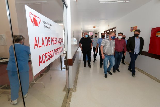 Profissionais de saúde caminhando pelo corredor do hospital em Rio Negrinho, com porta sinalizada para a ala de precaução à esquerda, durante preparação para abertura de 10 novos leitos de UTI COVID-19.