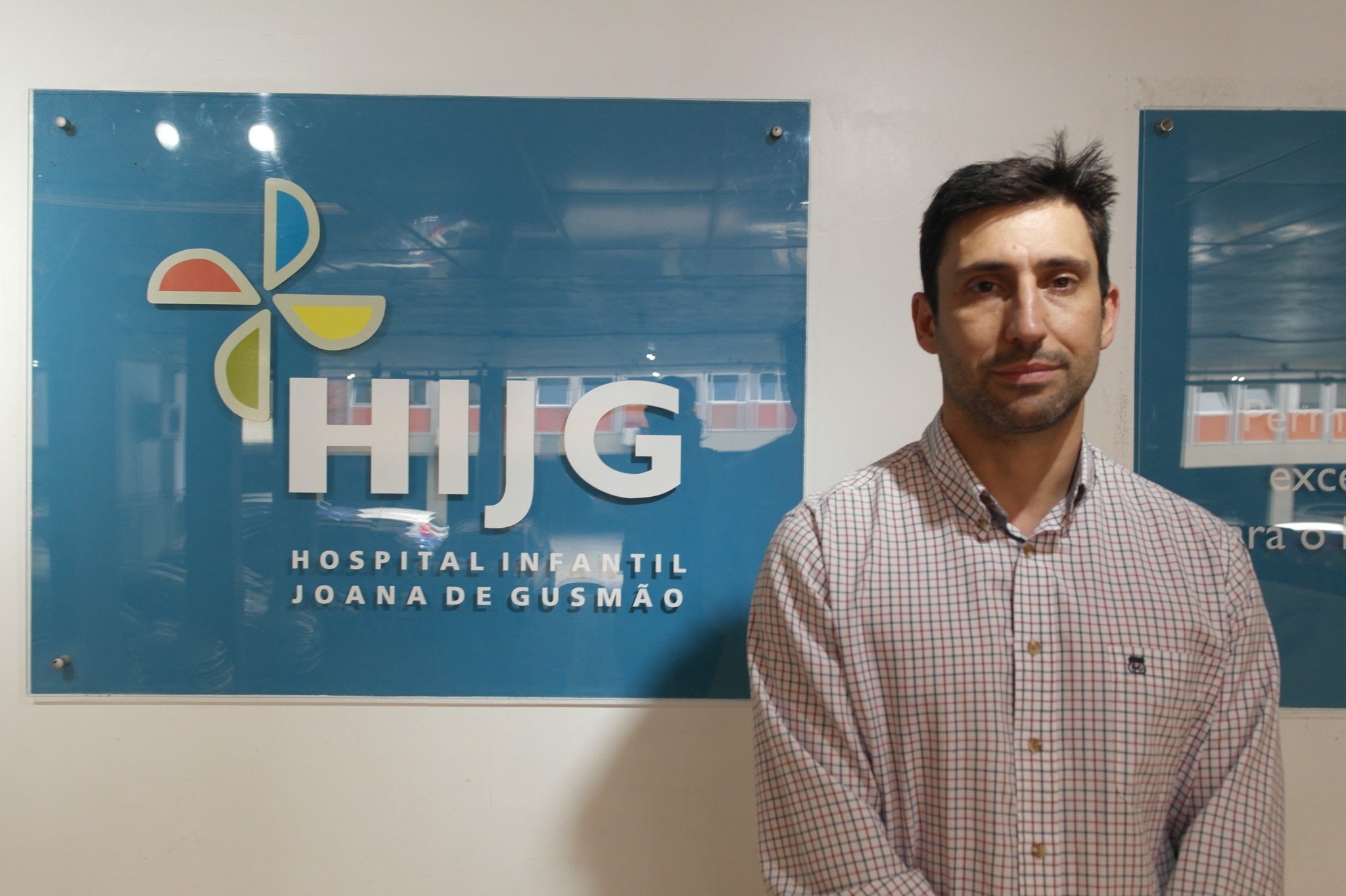 Homem em frente a painel do Hospital Infantil Joana de Gusmão (HIJG), em Florianópolis, SC. Ele veste camisa xadrez clara e posa para a foto ao lado do logotipo do hospital.
