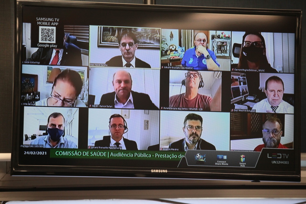 Tela de televisão exibe uma videoconferência com 13 participantes, todos com trajes formais ou de trabalho, alguns usando fones de ouvido e outros com máscaras. A reunião, datada de 24/02/2021, é uma audiência pública da Comissão de Saúde, conforme indicado na faixa verde na parte inferior da tela. O evento está relacionado à prestação de contas na área da saúde.