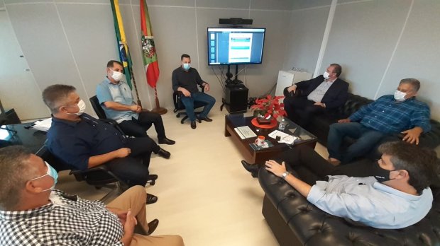 Sete homens, todos usando máscaras, participam de uma reunião em uma sala com sofás e bandeiras do Brasil e de Santa Catarina ao fundo. Um monitor exibe informações em uma apresentação digital.