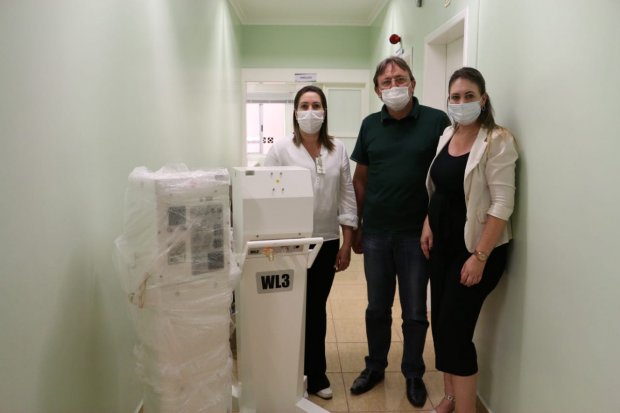 Três pessoas com máscaras de proteção posam em um corredor de hospital ao lado de um equipamento médico embalado. Duas mulheres e um homem estão em pé, próximos ao aparelho identificado como WL3.