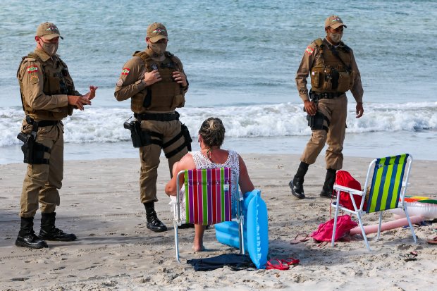 Policiais militares abordam uma mulher sentada em uma cadeira de praia, de frente para o mar, em uma faixa de areia. A mulher usa máscara e está cercada por objetos de praia como boias e toalhas. Três policiais, também de máscara, observam e conversam com ela. Ao fundo, o mar e a linha das ondas completam a cena de fiscalização sanitária.