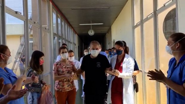 Paciente idoso usando máscara é aplaudido por profissionais de saúde ao sair de um corredor hospitalar, acompanhado por duas pessoas e cercado por aplausos de equipe médica em ambos os lados.
