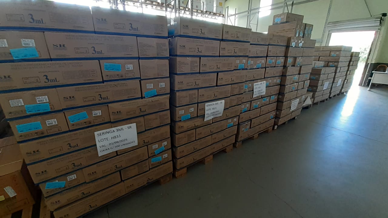 Imagem de um depósito com grandes pilhas de caixas de papelão empilhadas sobre pallets de madeira. As caixas estão organizadas lado a lado e identificadas com rótulos e etiquetas azuis.